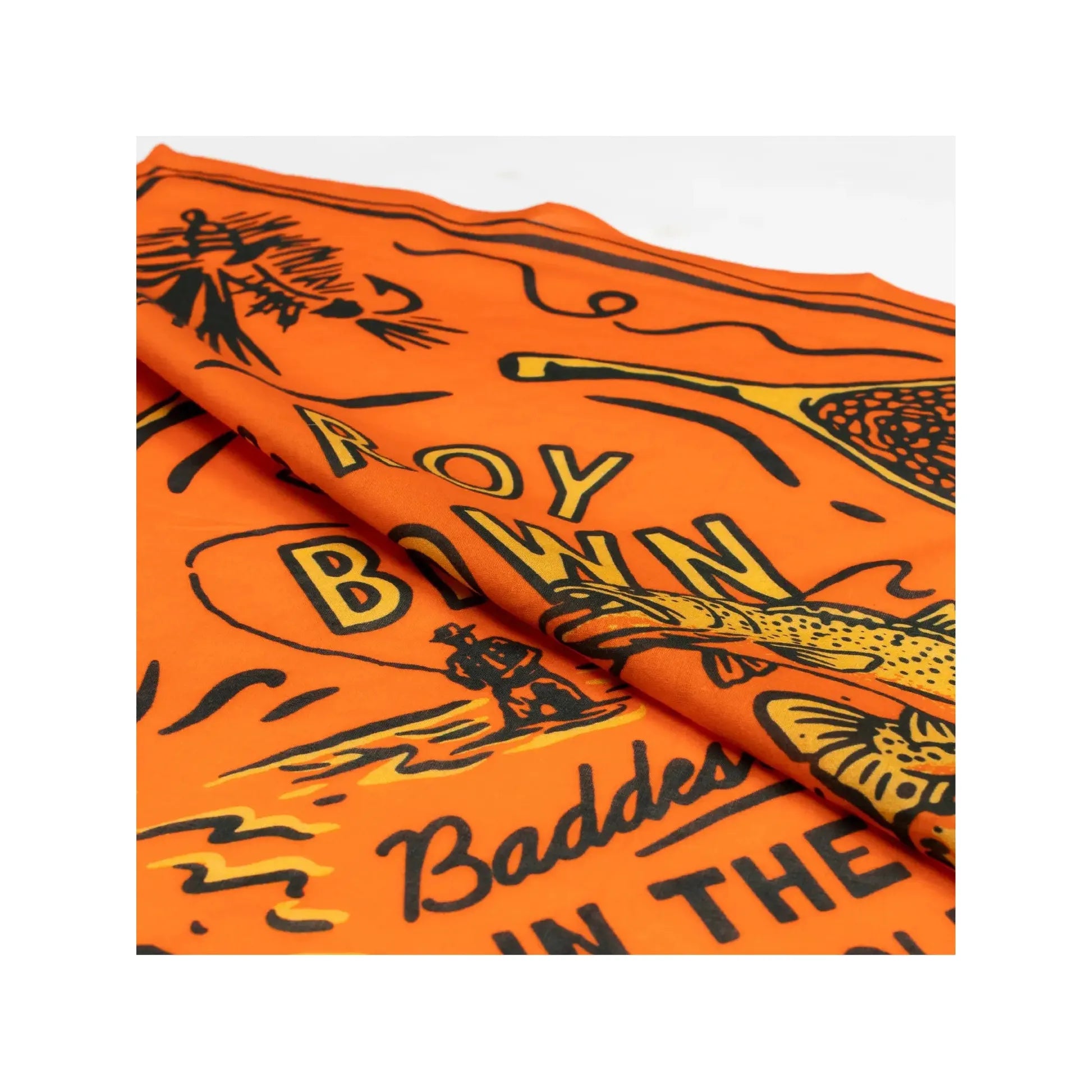 Close-up of an orange fabric with black illustrations and partially visible text reading "ROY BOWN" and "Baddest in the...". This Sendero Provisions Le Roy Brown Bandana is made from Organic Japanese Cotton, adding a touch of luxury to its striking design.
