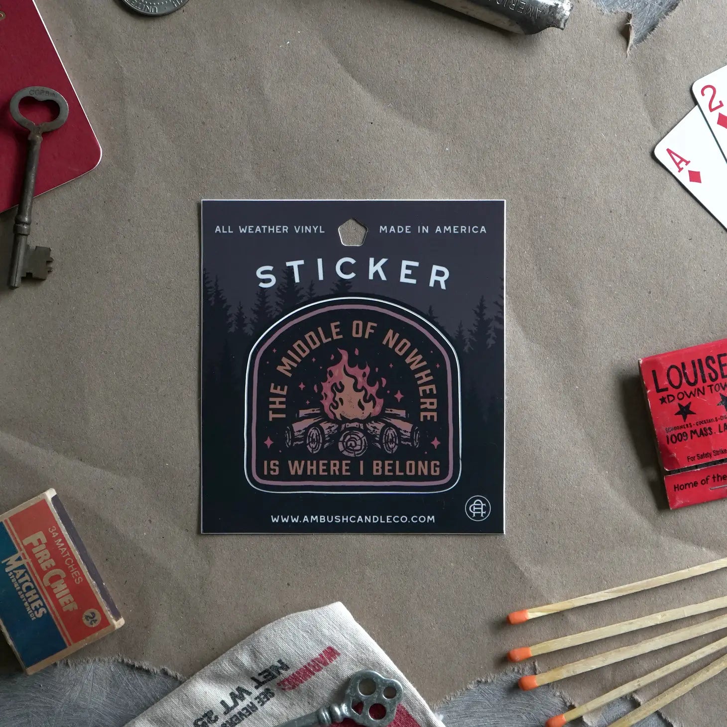 A weatherproof vinyl sticker from Ambush Candle Co, featuring the text "The Middle of No-Where is Where I Belong" and a campfire design, placed on a bed of fallen leaves.