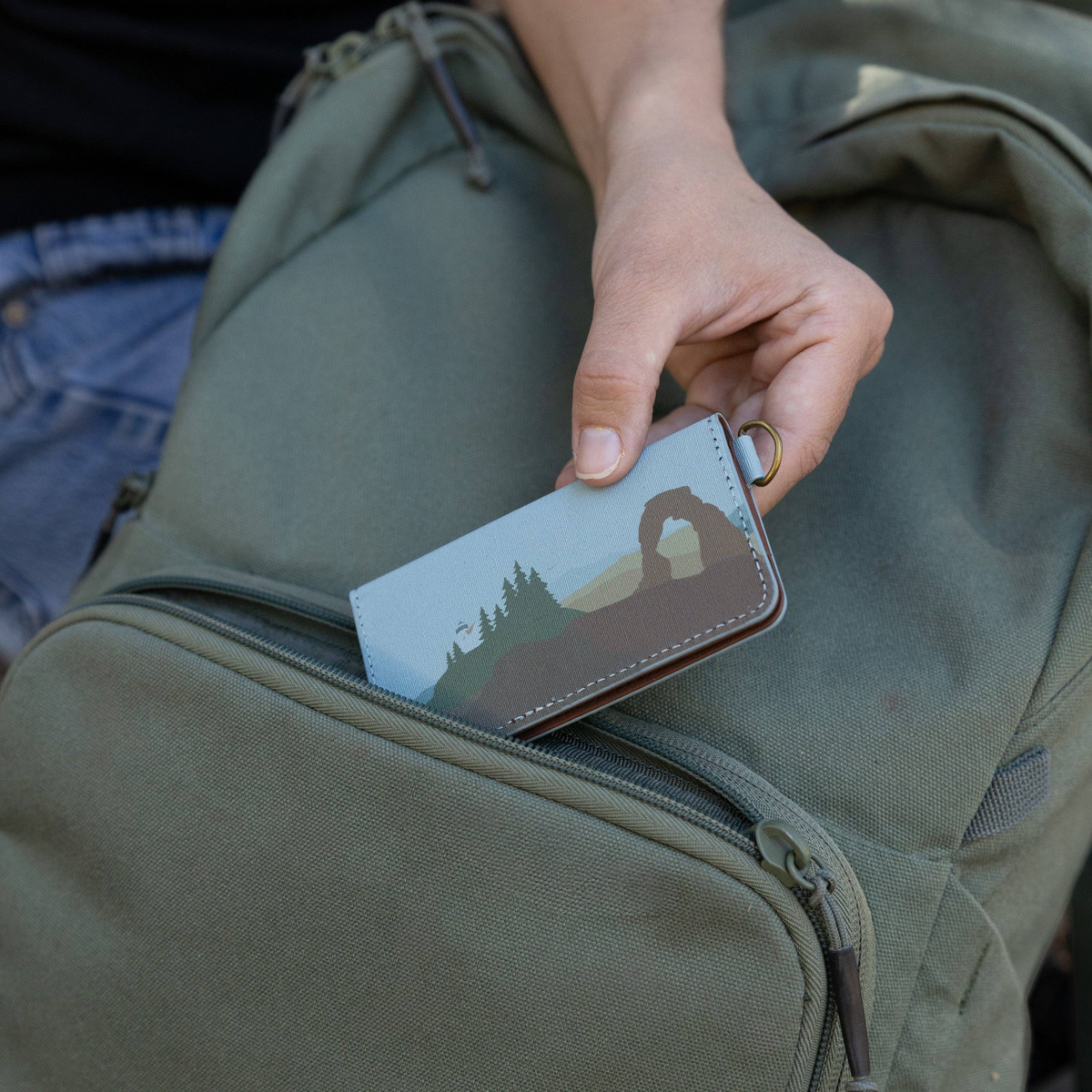 Forager Provisions LTD Wildtree card holder with Arches National Park design being placed into an olive green backpack.