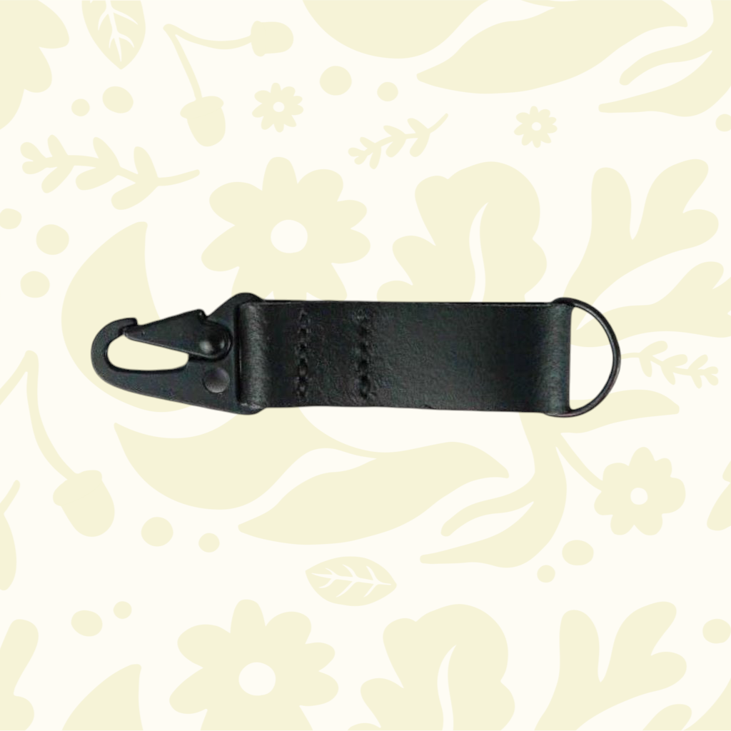 Kjøtt Forager Provisions black leather key fob with metal clip and ring, displayed on a cream floral-patterned background.