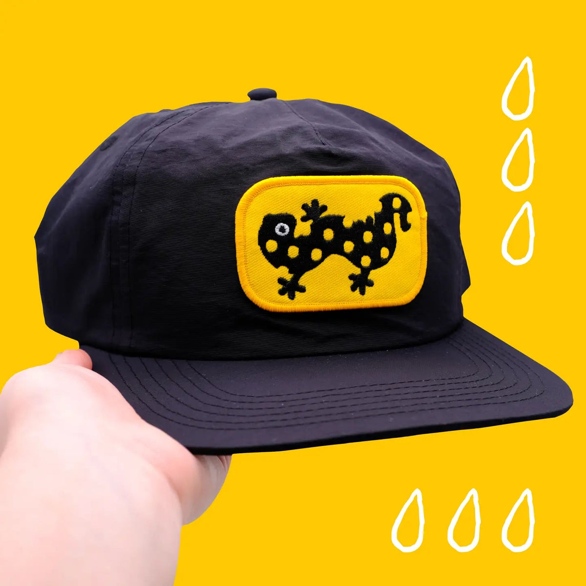 Hand displays Camp Mustelid Spotted Salamander Patch Cap on yellow background with illustrated raindrops, trendy hat fashion.