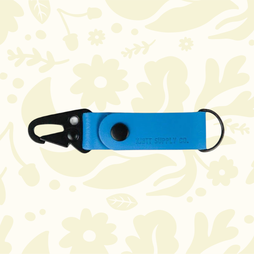 Blue Kjøtt Forager Provisions Leather Key Fob with black clip and ring, shown on a beige floral background.