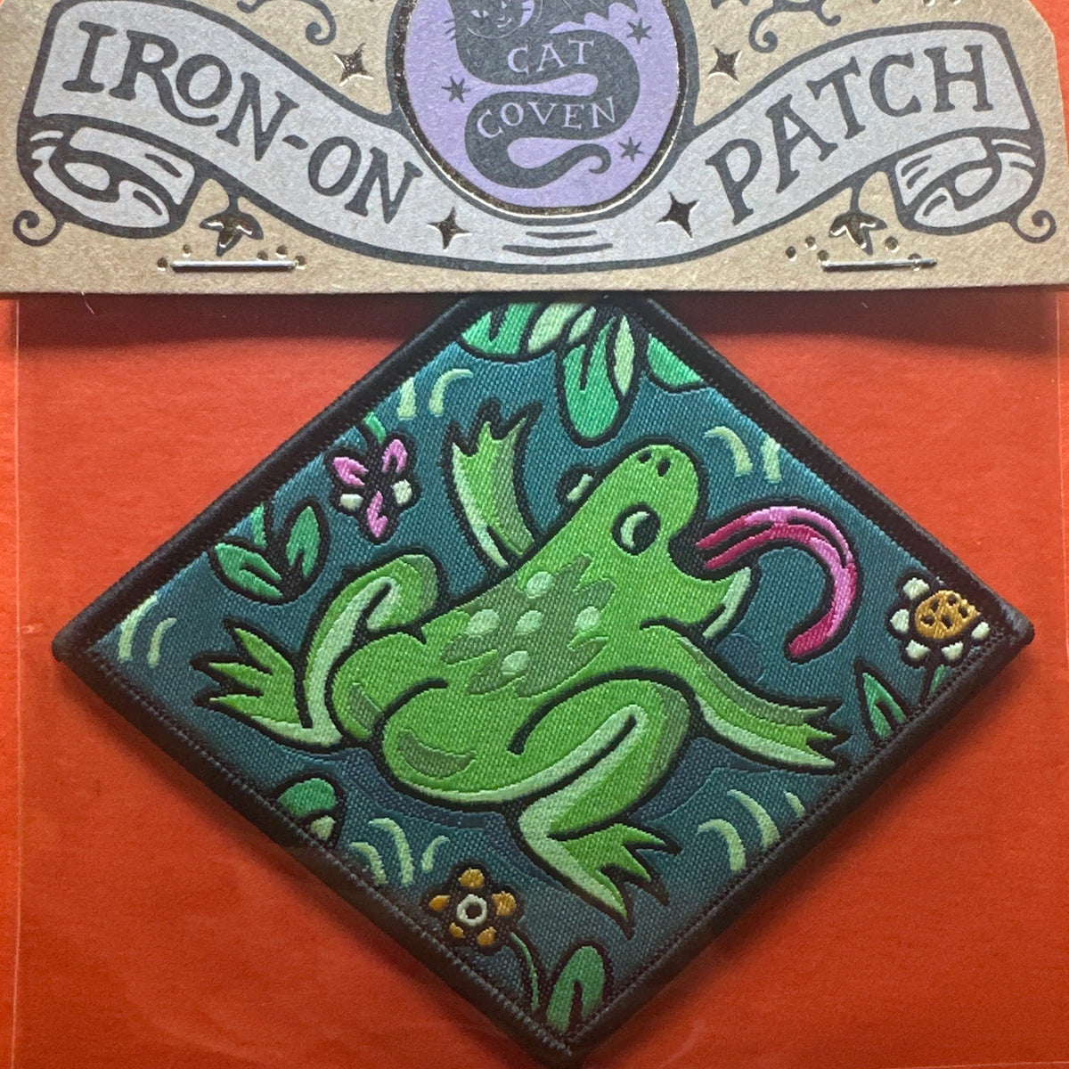 Forager Provisions LTD Cat Coven woven patch featuring a green frog with a pink tongue on a teal diamond background.