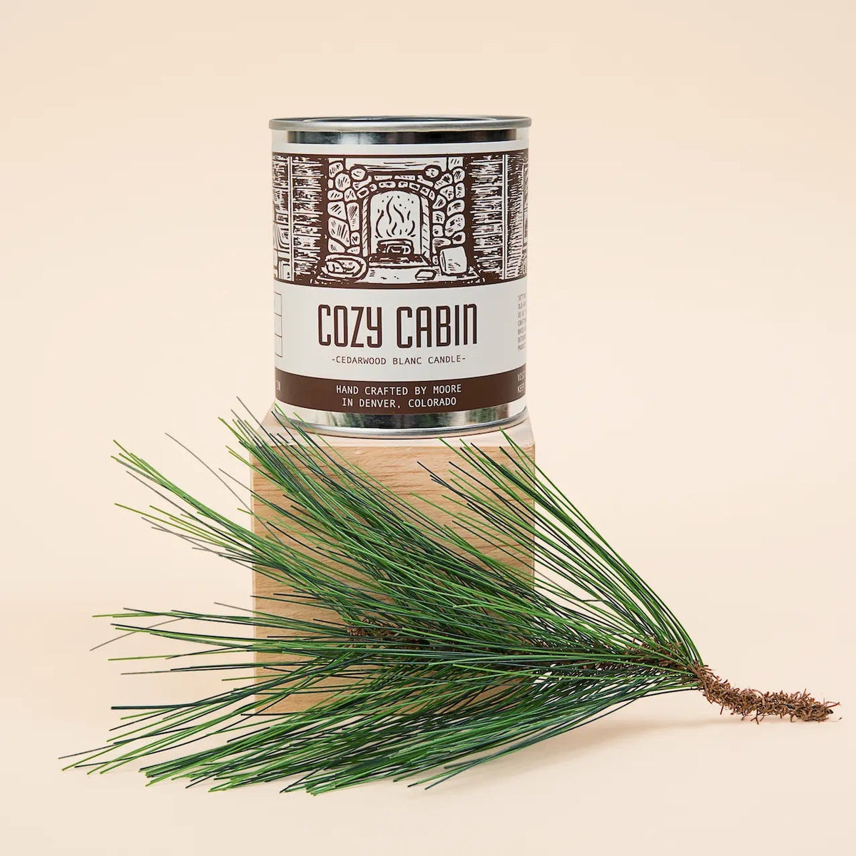 Forager Provisions LTD Moore Collection Cozy Cabin soy candle in a metal tin with a pine branch on a wooden block.