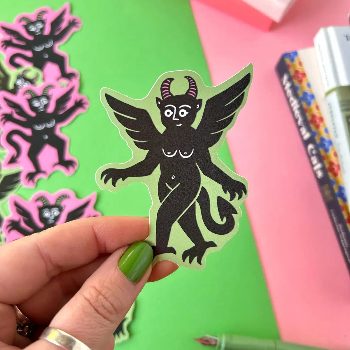Hand displays Stacey McEvoy Caunt Green Devil Medieval Vinyl Sticker on pink background, unique sticker art for laptops, journals.