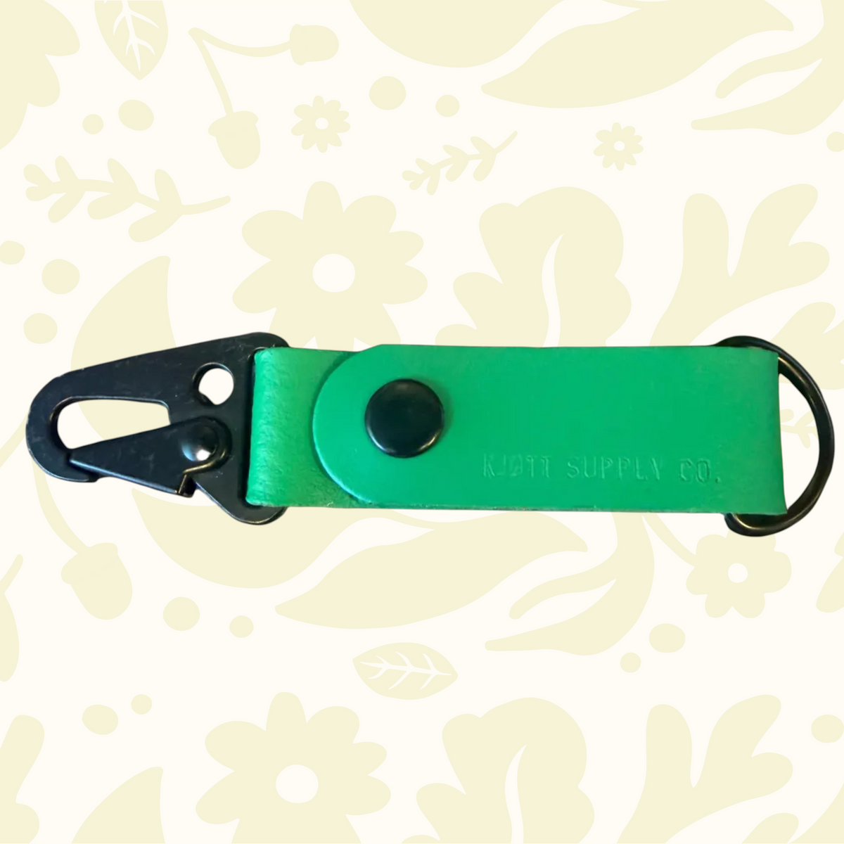 Green Kjøtt Supply Co leather key fob with clip, ring, and embossed logo on a floral background; luxury accessory.