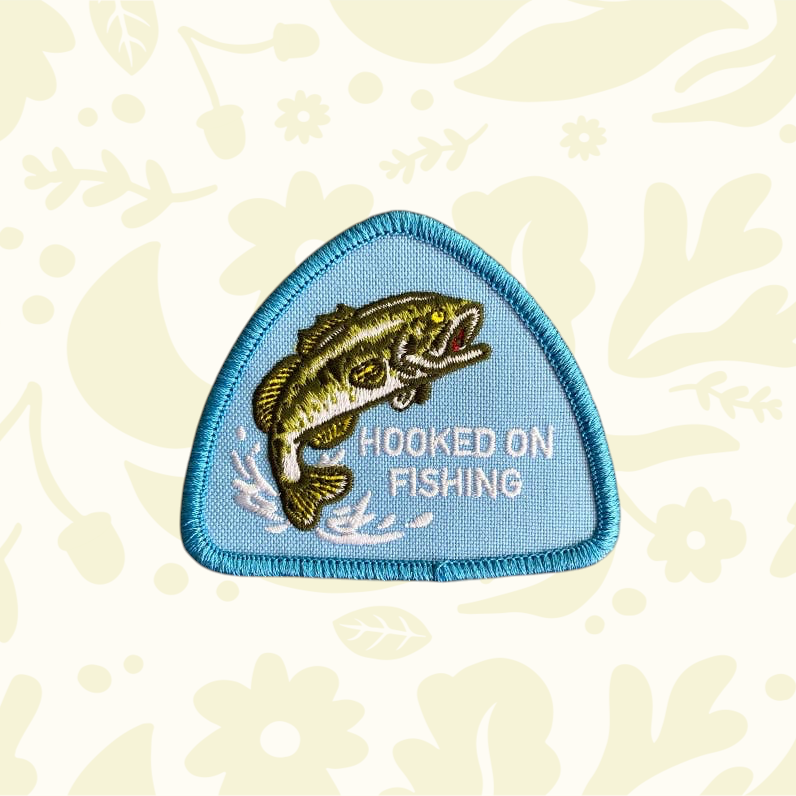 Forager Provisions LTD Hooked on Fishing triangular iron-on patch with a green bass fish on a light blue background.