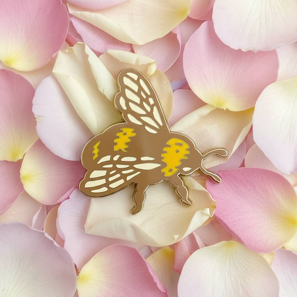 Forager Provisions LTD mustard and cream bee hard enamel pin with a mirrored finish resting on soft pink rose petals.