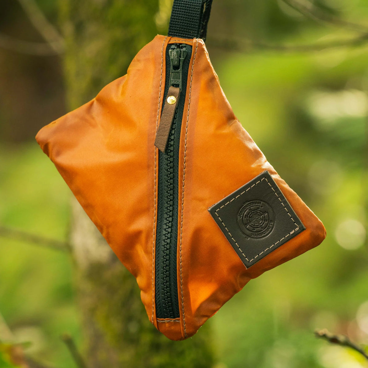 Forager Provisions LTD Journeyman Handcraft orange waxed canvas X-Pouch with a diagonal zip closure hanging outdoors.