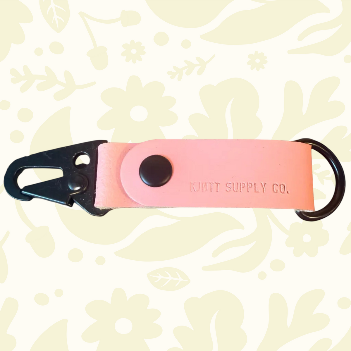 Pink Kjøtt Supply Co. leather key fob, black metal clip, embossed logo, floral backdrop. Premium accessory keychain.