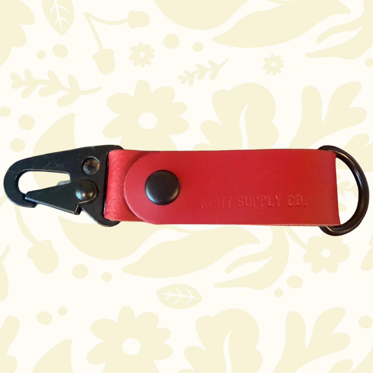 Red leather Kjøtt key fob with metal clip on floral fabric. Text: Kjøtt Supply Co Forager Provisions. Premium accessory.