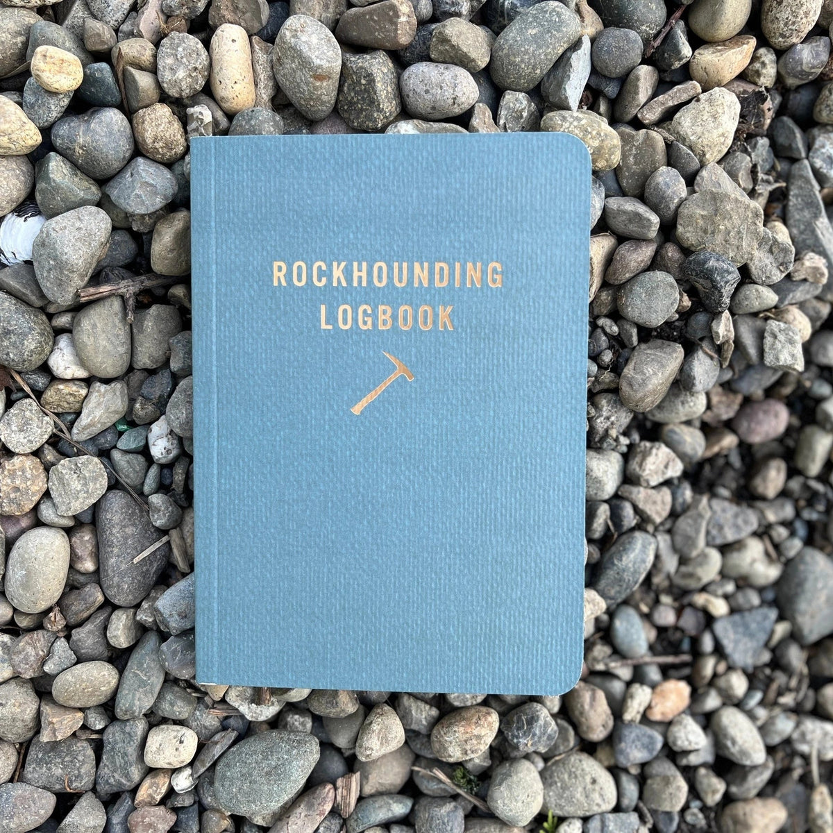 Forager Provisions LTD Mountaineer Books blue rockhounding logbook with gold foil text resting on a bed of river stones.