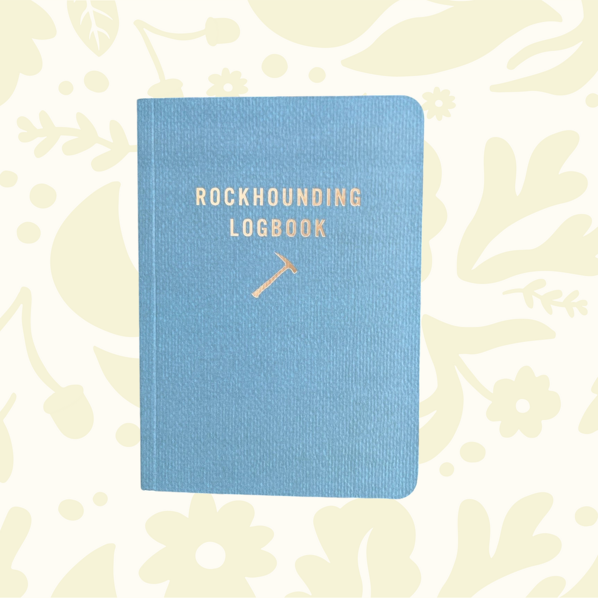 Forager Provisions LTD Mountaineer Books blue rockhounding logbook with gold foil lettering on a rocky ground surface.