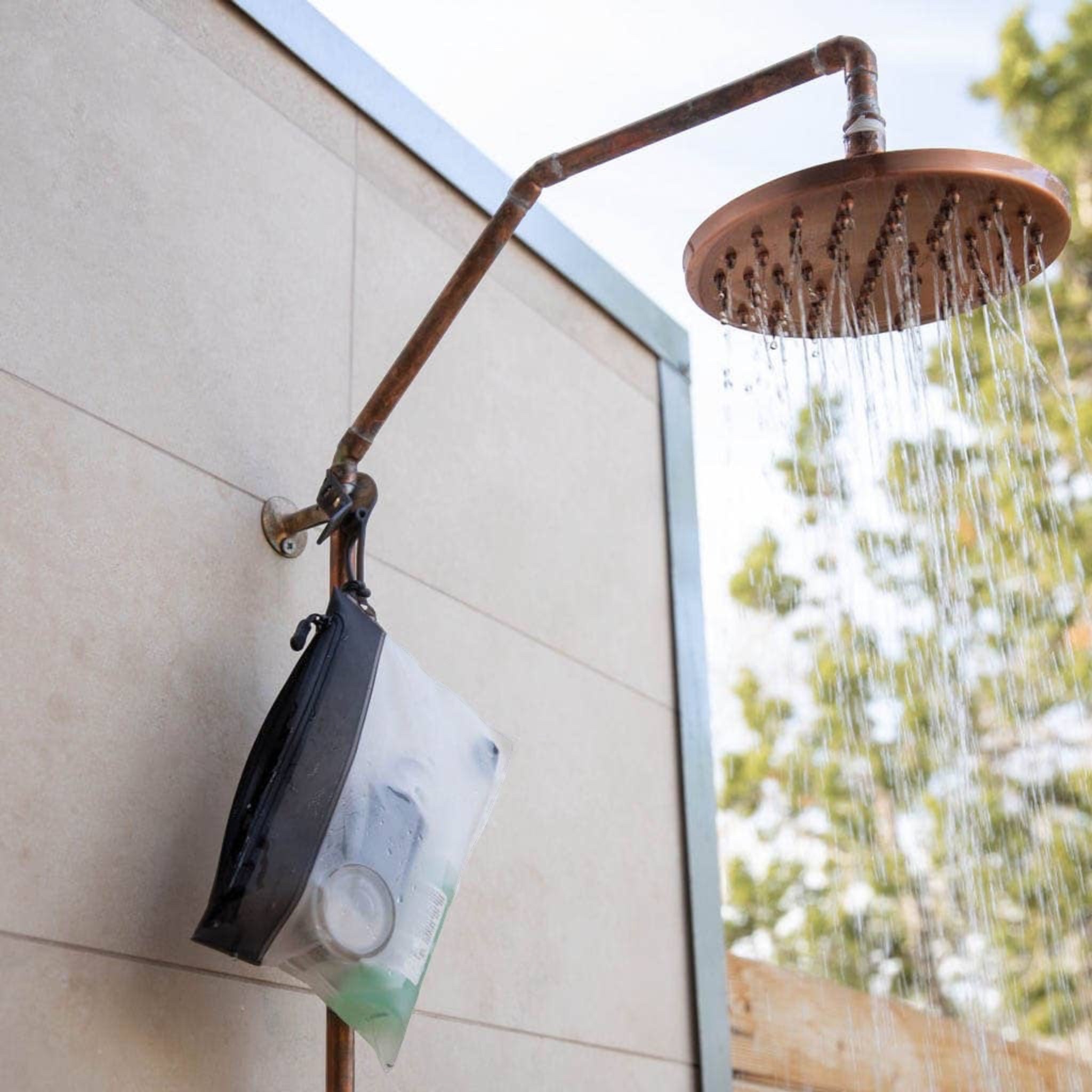 Forager Provisions LTD TSA bag hanging from an outdoor copper shower head, showing clear material and black zipper.