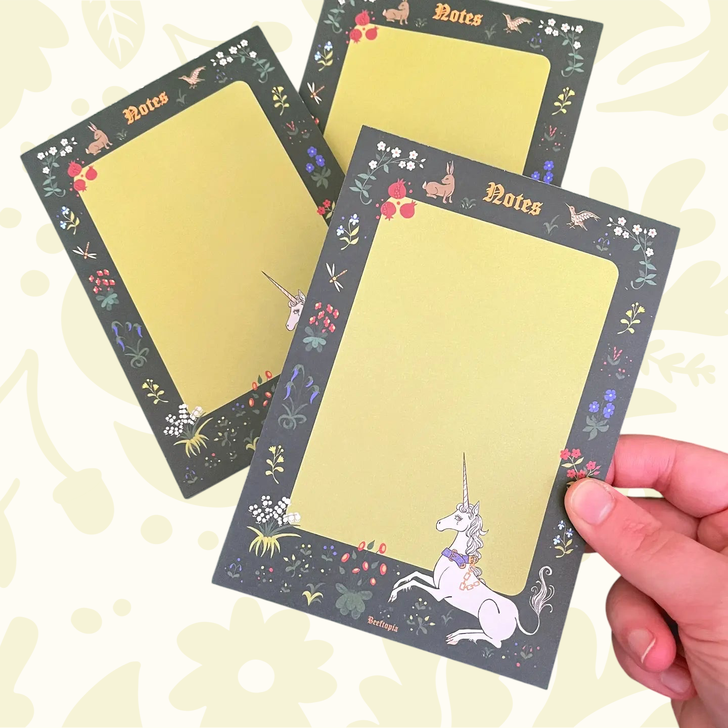 Hand displays Beeftopia Unicorn Tapestry Medieval A6 Notepad; two more on unicorn bordered, floral background stationery.