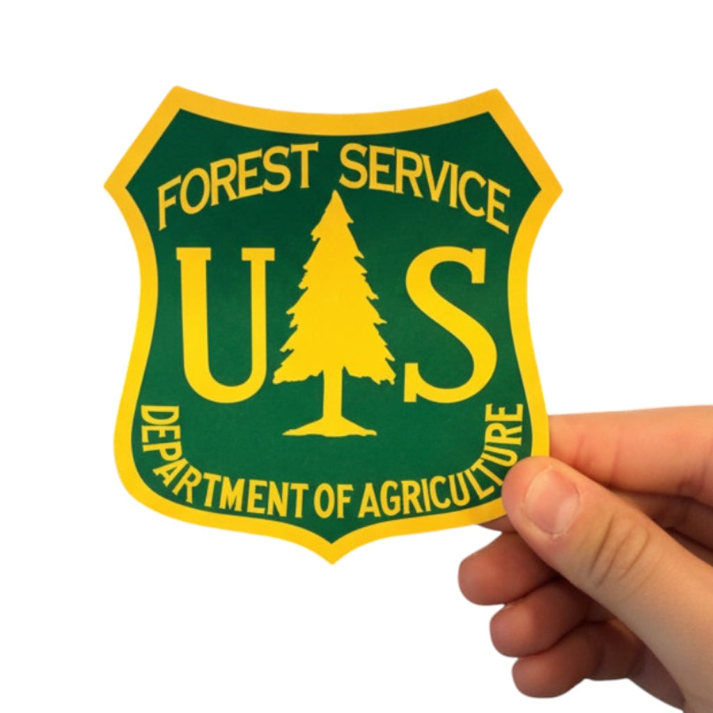 Hand holding Sawmill Shop’s green & yellow 5x3” US Forest Service Wood Shield sticker, made in the USA.
