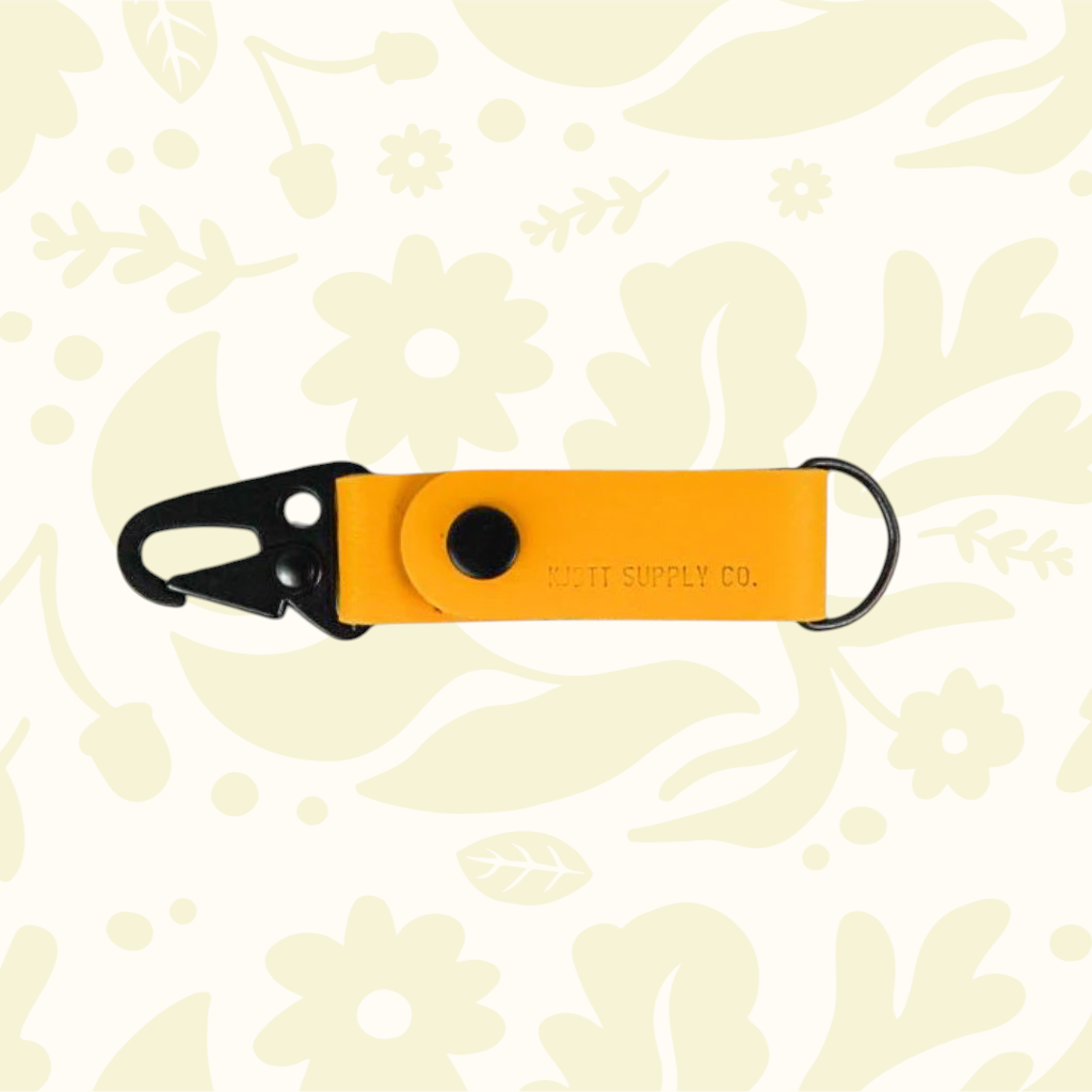 Kjøtt Forager Provisions yellow leather key fob with black metal clip, displayed on a floral patterned backdrop.