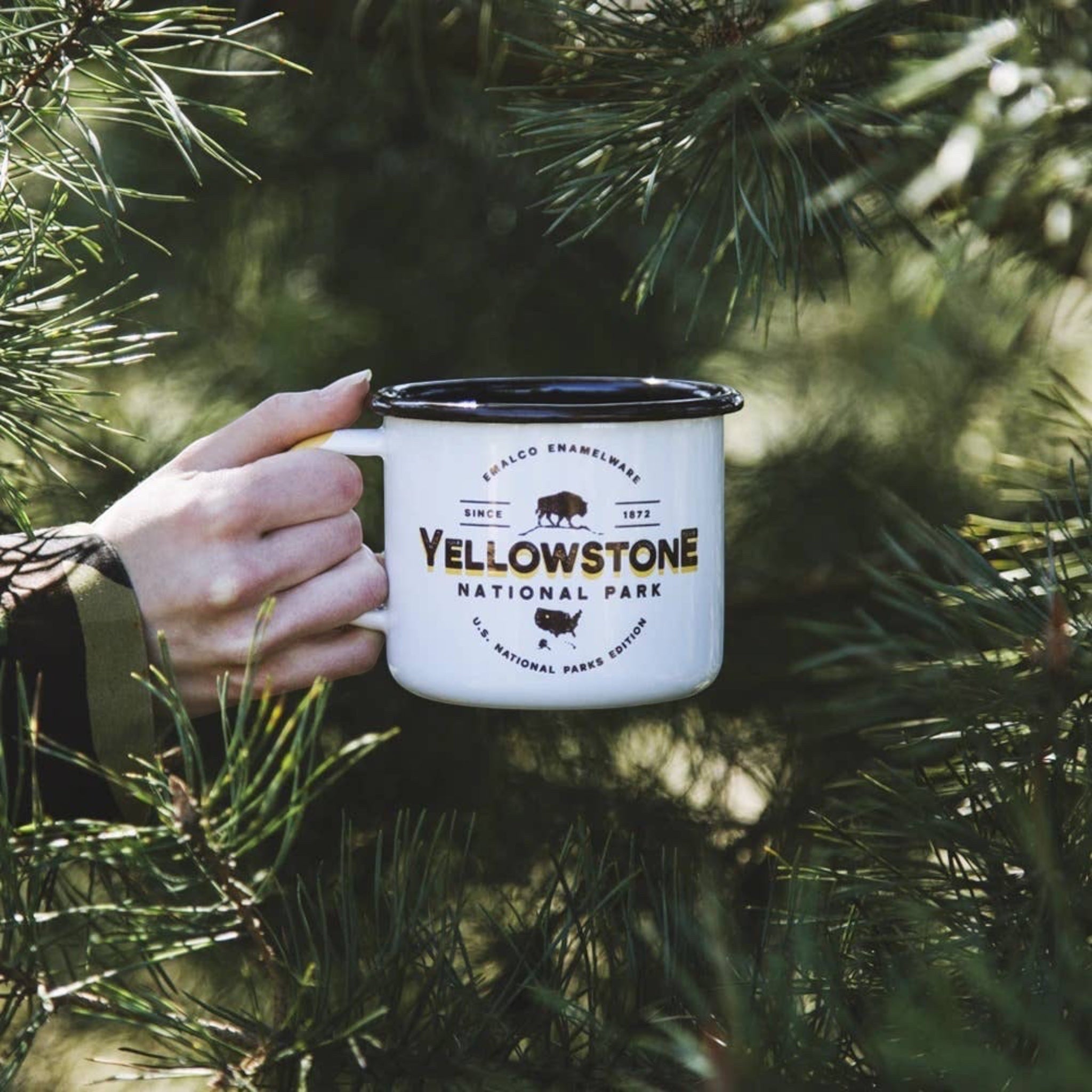 Forager Provisions LTD white enamel camping mug featuring a Yellowstone National Park graphic held by a hand.