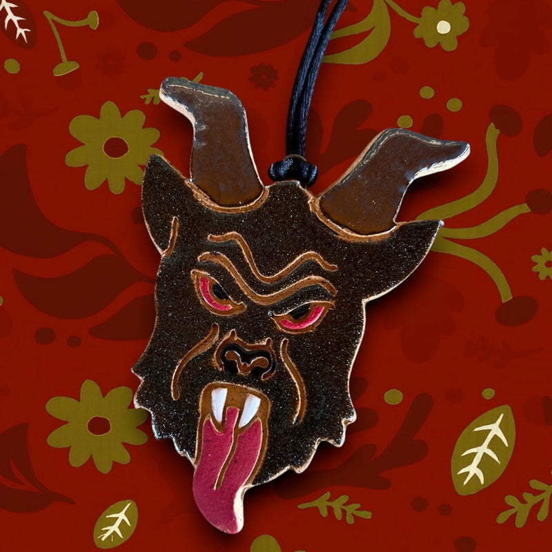 Horned creature head pendant with fangs and red tongue on black cord, displayed on a vivid red floral patterned background.