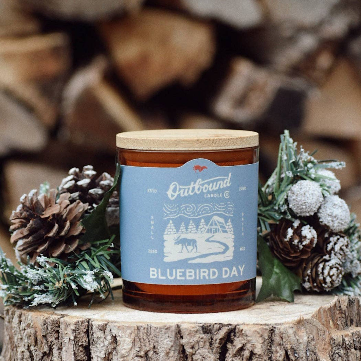 Outbound Candle Co. Bluebird Day soy candle in an amber glass jar with a bamboo lid, surrounded by pine cones.
